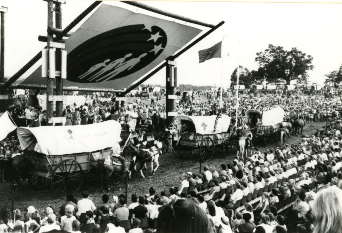 Wide shot of dozens of covered wagons and horse teams camped together in an open field at Valley Forge, Pennsylvania in July 1976, showing the scale of the Bicentennial Wagon Train Pilgrimage encampment