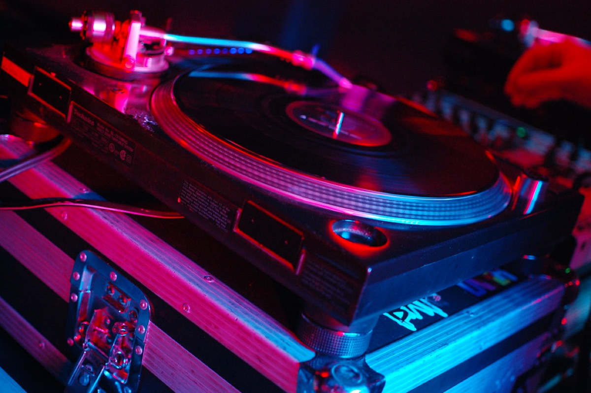 A vinyl record turning on a turntable in a dark room, a DJ's hand resting on the mixer alongside it