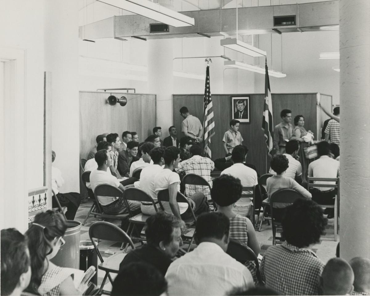 Archival black-and-white or period photo of Cuban refugees lined up outside or inside the Freedom Tower / Cuban Assistance Center in Miami during the 1960s or early 1970s, showing families waiting for processing