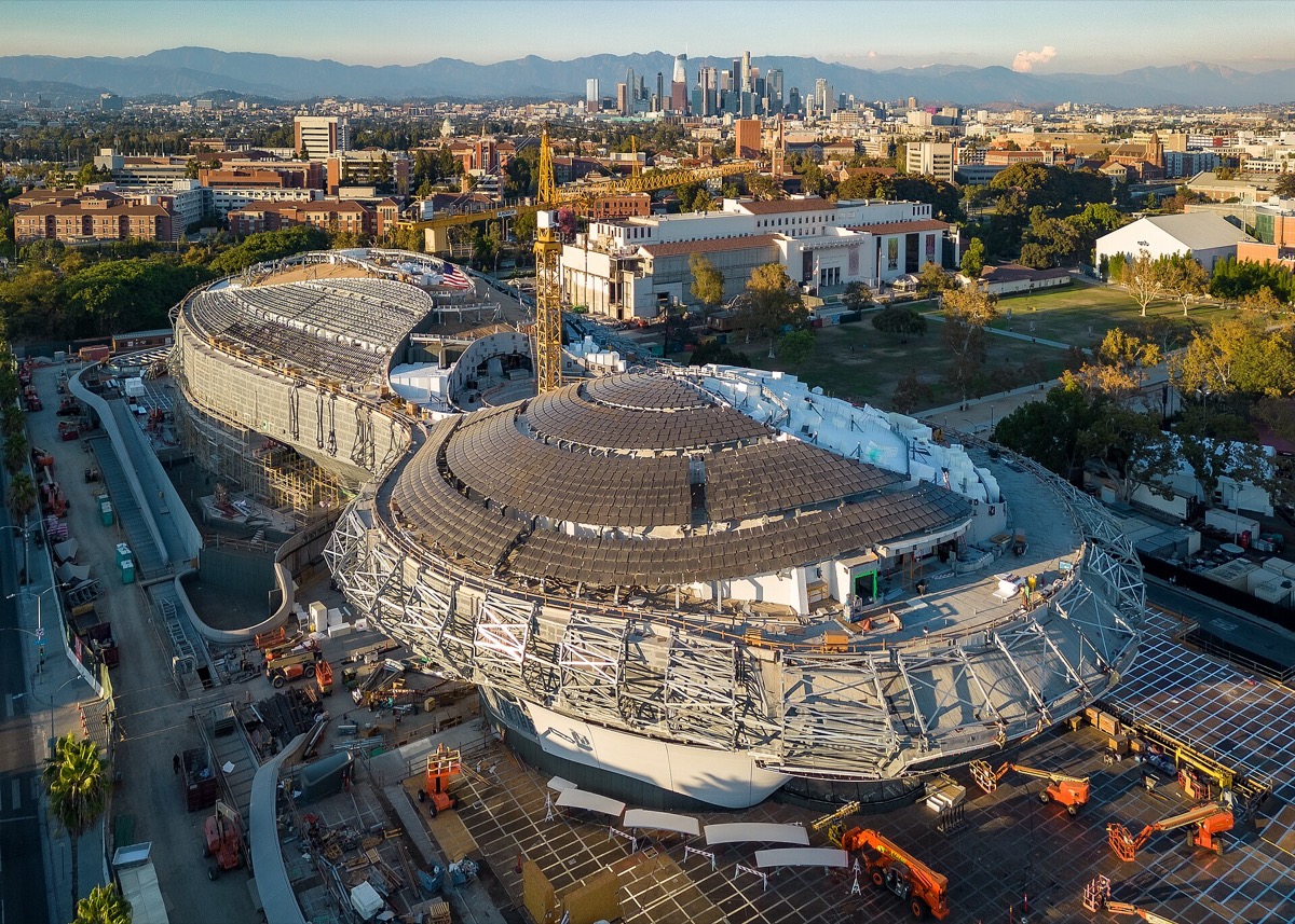 The Lucas Museum of Narrative Art under construction in Exposition Park, Los Angeles, its curving spaceship-shaped roof rising above the park with the downtown skyline behind