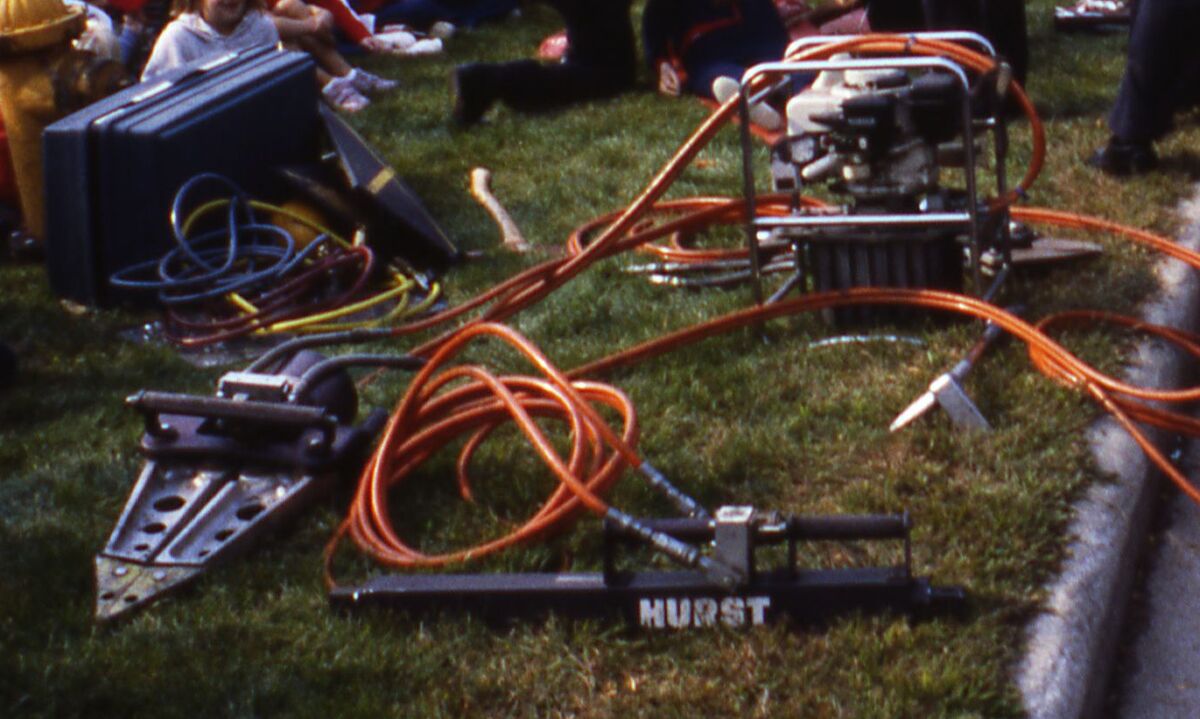 An early Hurst rescue tool set laid out on the grass at a 1981 fire department demonstration in Columbus, Ohio - a black hydraulic ram with HURST stamped along its body, a pair of spreader arms, coiled orange hydraulic hoses and a petrol driven pump