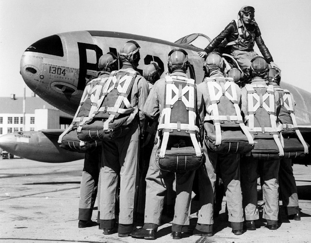 USAF flight instructor briefing Aviation Cadets in parachute harnesses beside a Lockheed F-80 Shooting Star jet, 1940s