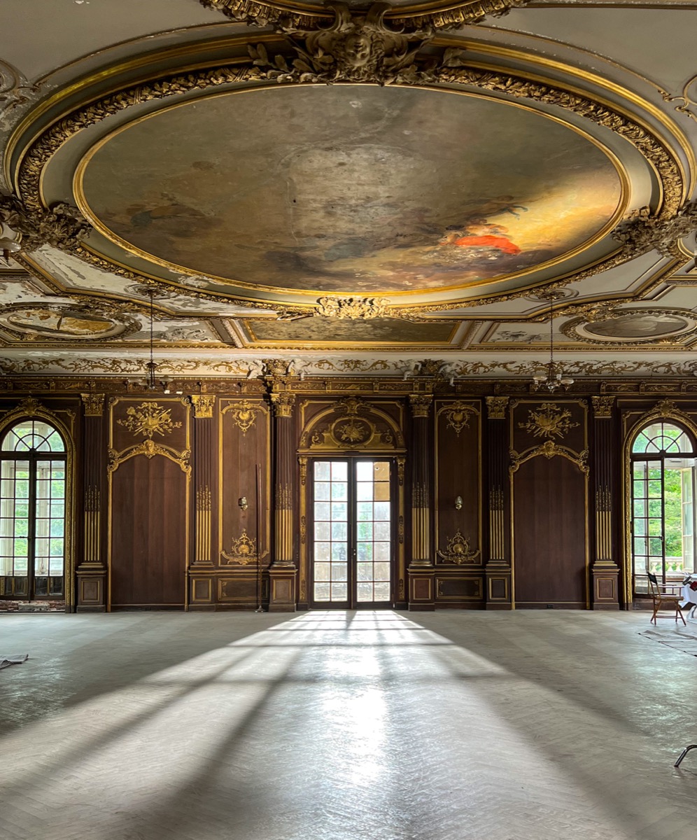 Wide interior view of the grand ballroom inside Lynnewood Hall looking east, showing the ornate gilded ceiling, tall windows and marble floor of the restored Gilded Age room
