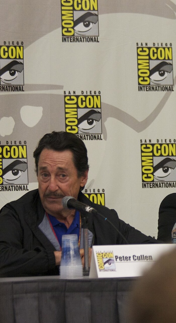 Peter Cullen, older man with grey hair and beard, speaking into a microphone on stage at a Transformers fan convention panel
