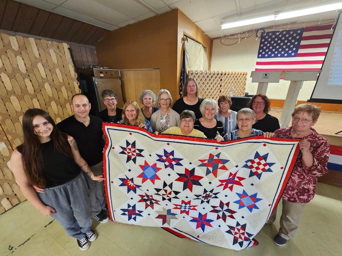 A group of volunteer quilters, mostly women of varying ages, sitting at sewing machines and long fabric-cutting tables in a community hall or church basement, working on red-white-and-blue patriotic quilts, USA/veteran quilting bee, wide group shot