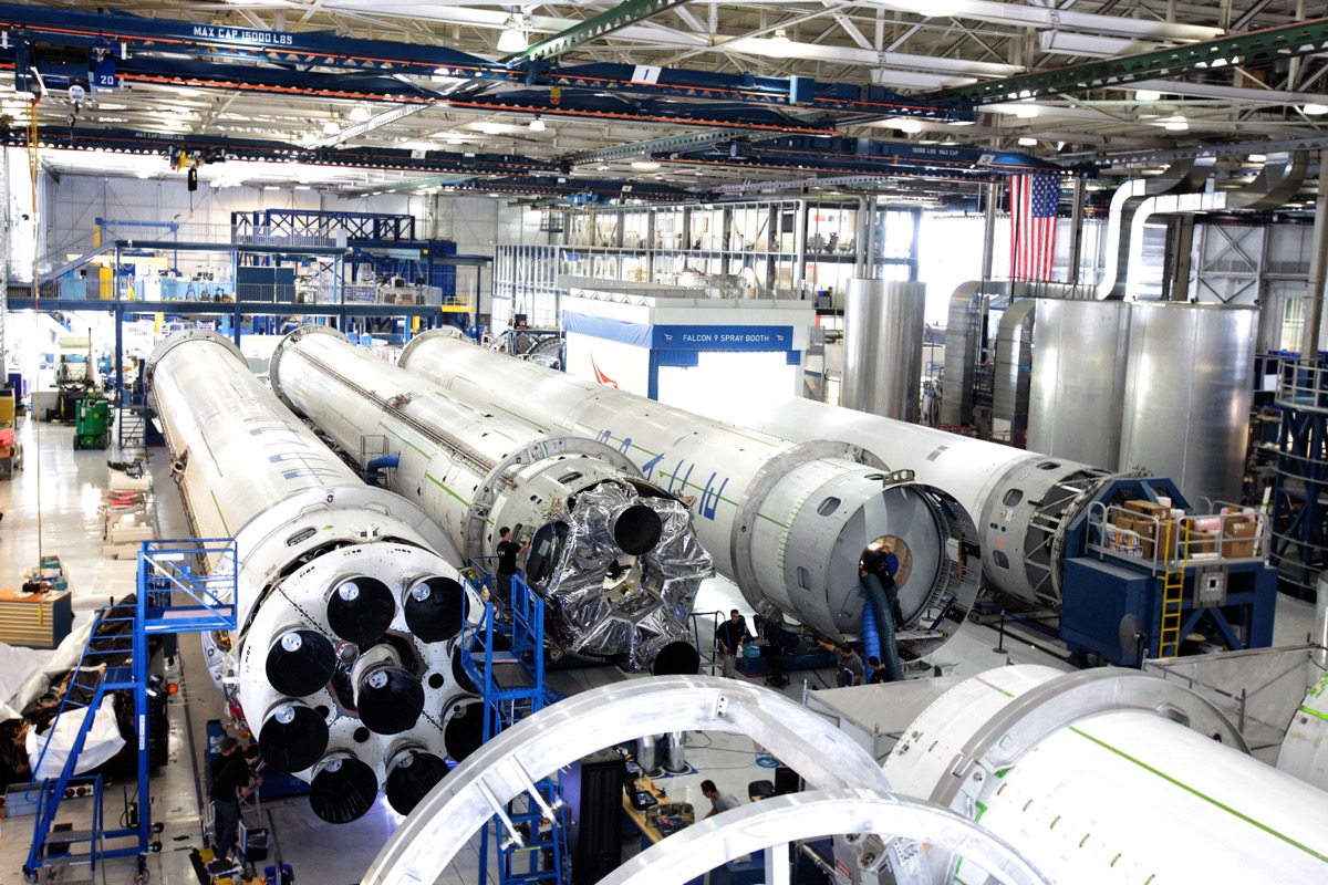 Wide interior view of the SpaceX rocket factory floor in Hawthorne, California, showing technicians and machinists in work clothes assembling Falcon 9 boosters or Merlin engines among tooling and hardware
