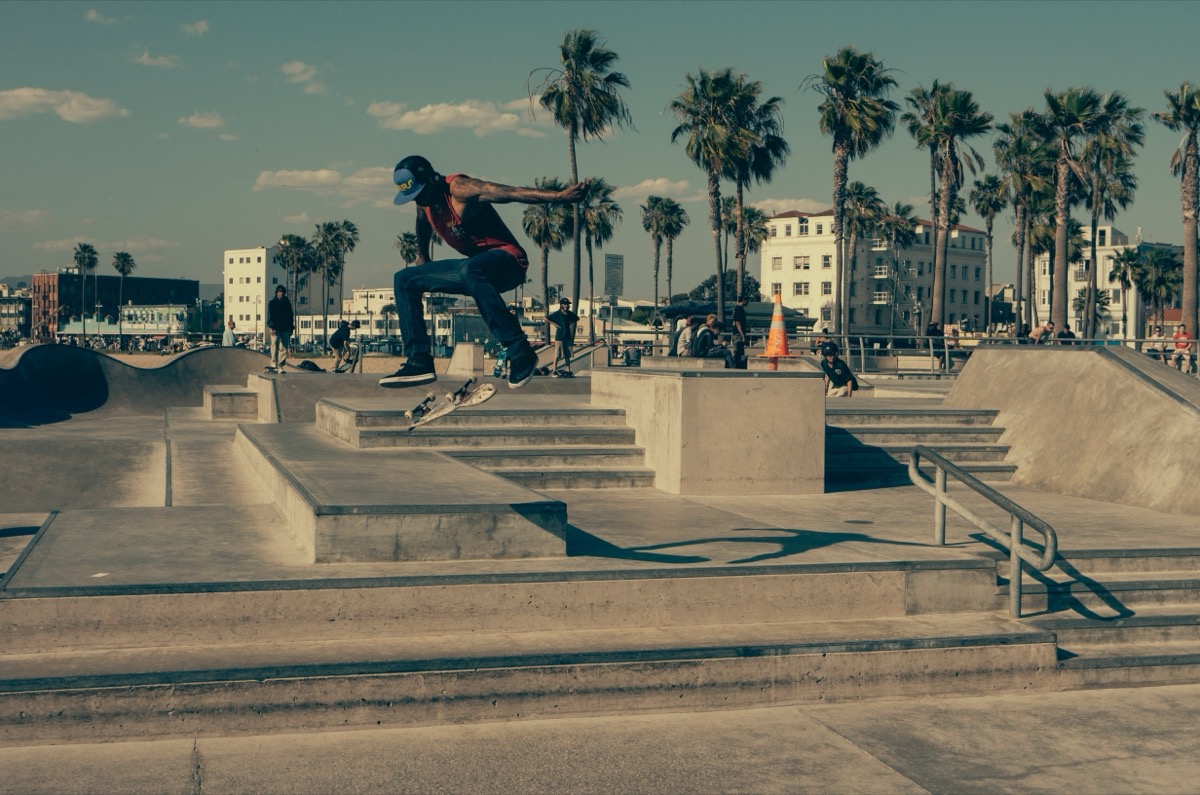 Kids and teenagers skating at a sunny outdoor public skatepark, multiple skaters visible on ramps and bowls, community park setting, no single person as the main subject